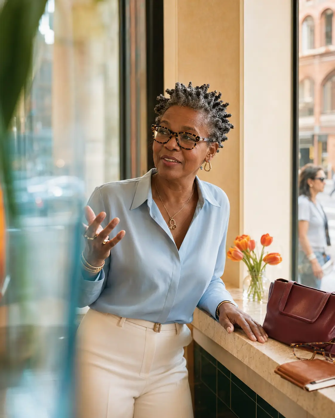 A well-dressed midlife woman speaking warmly beside a sunlit New York cafe window.